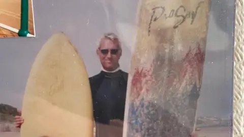 Louis Thomas-Hudson A old retro-styled picture of Ronnie Hudson with two long white surfboards, holding on either side of him. The gentleman is wearied a black wet suit, he has black sunglasses on, with grey short-ish hair.