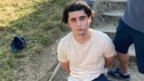 Daniel Khalife, wearing a white t-shirt, is pictured sitting on the ground outside with his hands behind his back after his arrest
