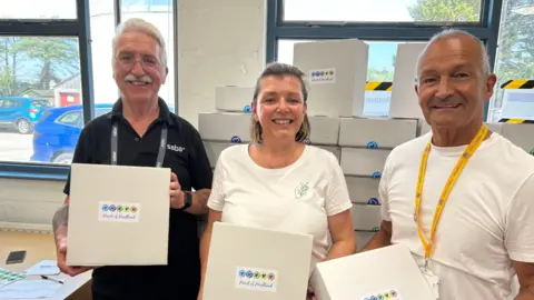 Photo shows volunteers standing together smiling at the camera. Glynn Pettifer is wearing a black T-shirt while Kelly Hallett and Pete Eddy are both wearing white T-shirts. They are holding boxes. Behind them are stacks of boxes and large windows.