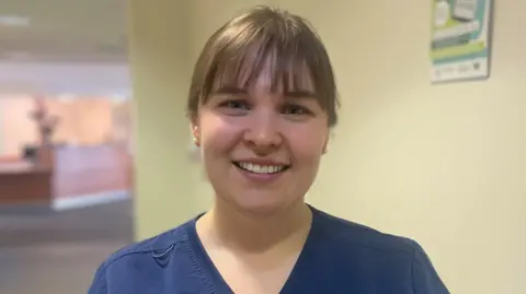 Dr Samantha McCarthy standing in a corridor with yellow walls, looking at the camera. She is wearing dark blue scrubs and has her hair tied back