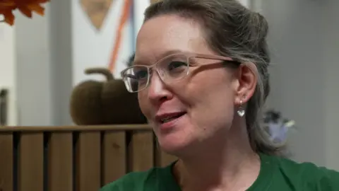 Rebecca Goodier has light brown hair which is tied up and is wearing light pink framed glasses and a green t-shirt and a heart shaped pendant earring.