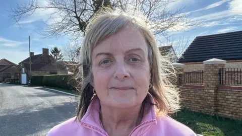 Fiona Taylor is wearing a pink jacket. She has medium length hair which is blonde at the top and browner towards the ends. Behind her is a road and houses.