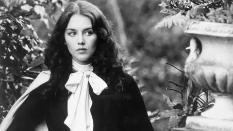 A black and white image of a woman with brunette hair in a garden with stone ornaments. 