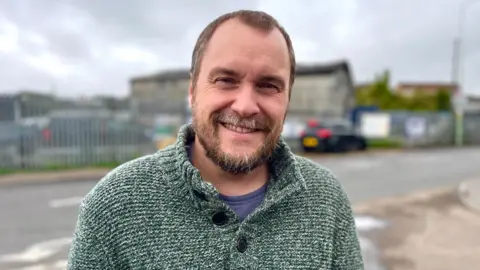 Martin Giles/BBC Philip Cook with short light brown hair and a short beard, wearing a white flecked fisherman-style grey jumper, with buttons at the neck and behind him, across a road, a warehouse