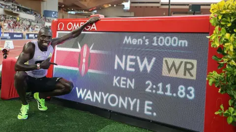 Emmanuel Wanyonyi poses for a photograph next to a digital screen showing his name and world record time after winning the 1,000m in Monaco