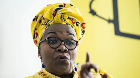 Alice Nkom wearing a yellow headscarf and shirt purses her lips and raises a finger as she speaks to an unseen interviewer.