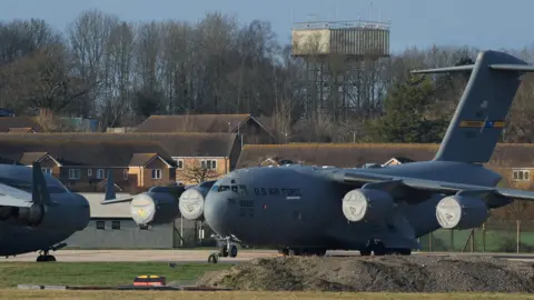 Reuters A large dark grey plane with US Air Force written on it is seen sitting on the tarmac at a small airport. Another plane of a similar size and colour is parked next to it, mostly out of shot. A number of houses can be seen in the background, as well as trees and a large water tower. 