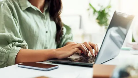 A woman with dark hair wearing a green button-up shirt with puffed three-quarter length sleeves. She is sitting at a white desk, typing on a grey laptop. In the background there is a plant out of focus.