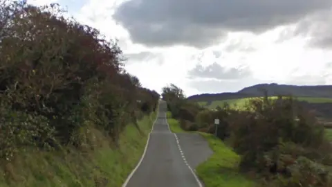 A long straight tarmacked road runs down the centre, with a high grass verge on the left adorned with trees. There is a lay-by on the right, and the road is also lined with grass and trees but lower down. It is a cloudy day and there are rolling green hills in the distance on the right.