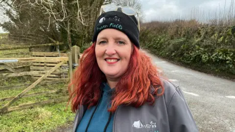 Helen Bantock standing next to field gate and lane covered with patches of frost. She has red hair and is wearing a grey jacket over a blue hoodie and a black hat with her business name Frowen Fields on it