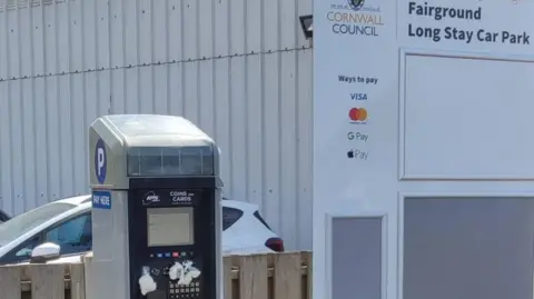 VIA LDRS A parking meter can be seen with a sign to the right which says Fairground Long Stay Car Park, with Cornwall Council branding and a white car can be seen in the background 