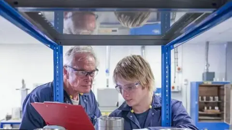 A man in a blue overall is holding a red clip board and wearing goggles next to a boy in blue overalls who is also wearing goggles.