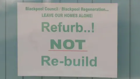 A white paper sign in a house window - it has the words Refurb NOT Re-build in green letters