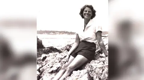 Se adjunta una fotografía en blanco y negro de una joven con una camisa blanca y pantalones cortos oscuros sentada sobre unas rocas.