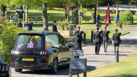 A funeral procession is passing through a narrow road surrounded by trees and heading towards a crematorium. A black funeral car can be seen with a wooden coffin inside with yellow flowers on the top of it. Members of the Royal British Legion dressed in black suits can be seen walking in front of it and carrying tall flags.