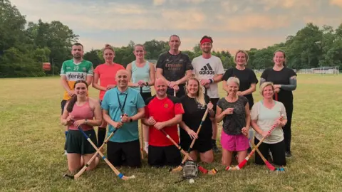 Camanachd Association A group of Camanachd players consisting of men and women stand on the photo and hold camans (sticks for playing the game). 6 people in the front row and 7 in the back row.