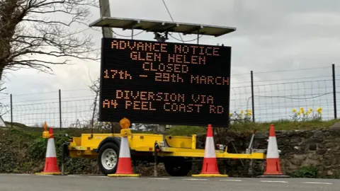 BBC A large digital sign on a yellow trailer at a roadside with four orange and white traffic cones in front of it. The sign reads: Advance notice, Glen Helen closed 17th - 19th March. Diversion via A4 Peel Coast Road.