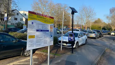 LDRS A car park with a board displaying payment options. Cars are parked in bays on a sunny day. There are a few trees around the car park.