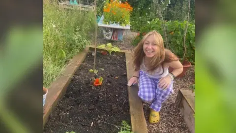 Laura Mathurin Laura is on one knee next to a rectangular piece of wood which is full of soil with a few plants in. She has blonde hair, a white top, and purple and white chequered trousers on. 