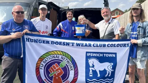 A line of three men and three women holding a white banner which says "Hunger doesn't wear club colours" and featuring the Ipswich Town logo and a FSF network logo (a red hand holding a green fork). One of the men is holding a blue collecting bucket above the banner. The open rear door of a van is behind them.