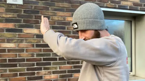 Justin Allison outside Newport Magistrates' Court, he wears a pale grey sweatshirt and a grey ribbed north face beanie hat, while holding his left arm up to partly cover his face. 