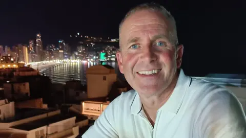 Les Howard, who is wearing a white polo shirt, is starring directly at the camera and is smiling. There is a city skyline in the background. 