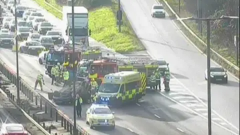 National Highways A motorway camera shows a number of emergency services vehicles. Traffic is backing up behind them. A car is on its roof. 