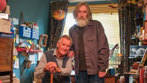 BBC/HOT OLIVES PRODUCTIONS LIMITED Pearce Quigley stands next Sir Michael, who is sitting on a chair while leaning on a walking stick. A range of items are displayed on the walls and shelves around them in a room.