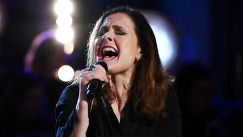 Alison Moyet sings into a microphone she is holding with her right hand. Her eyes are closed and her mouth is wide open. She has long brown hair and is wearing a black shirt.