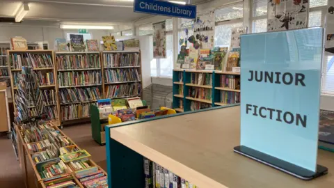 Children's section of a library with shelves of books with coloured spines. 