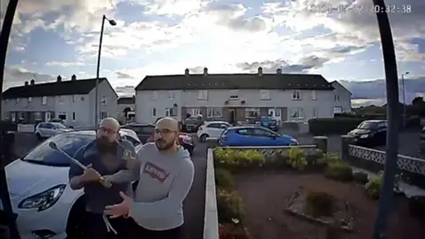 Crown Office Doorbell camera footage showing two bald men, both with glasses and beards. They both have their hands on some kind of metal pole. 