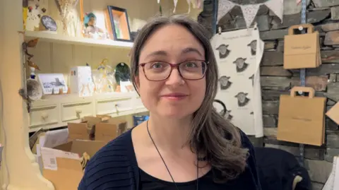 A woman with long brown hair, swept onto one side so it covers her shoulder. She's wearing red-brown classes and she's slightly smiling. In the background there's a tea towel hung on the wall with pictures of sheep on it.