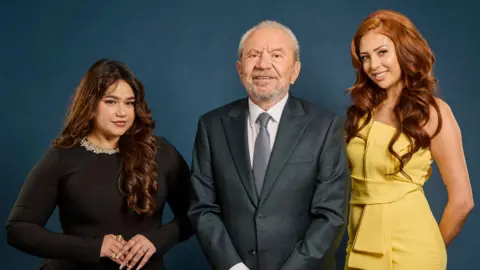 Three people pose for a studio photograph against a dark blue backdrop. Lord Alan Sugar stands in the centre wearing a dark suit and tie. Karishma Vijay stands to one side wearing a black dress, while finalist Pascha Myhill stands on the other side wearing a yellow sleeveless outfit.