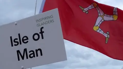A white board on a brown stick is held in the sky, it reads Isle of Man. Behind it a red Manx flag also flies against the backdrop of a cloudy sky. It bears a yellow and white triskelion in its centre.