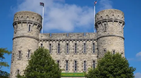 A view of the front of the former county barracks building, it is a pale grey building made of stone with two towers, with flags raised, and trees at the front 