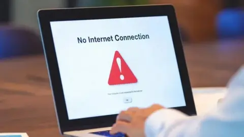 A stock image of laptop with no internet connection. The screen is open and the words no internet connection are written on the white screen, with a red triangle with an white explanation mark in its middle. A man's hands are resting on the keyboard.