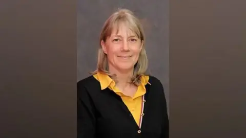 Dacorum Borough Council A studio-style portrait of Sally Symington with long blonde hair against a plain grey background. She is wearing a black cardigan over a yellow collared shirt and is facing the camera.