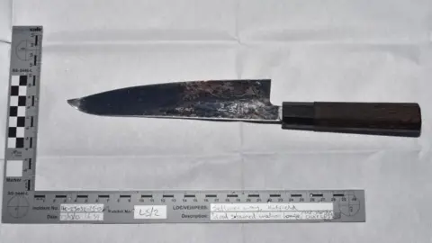 Hertfordshire Police A black and white image of a dark-handled kitchen knife. The blade has one straight edge and an edge which is wide at the base and slopes to a point. The point of the knife in bent up. The knife is framed by a L-shaped ruler which gives indications of measurements.
