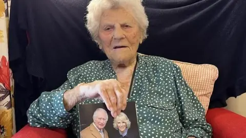 Ethel Caterham sat holding a card with a picture of the King and Queen on the cover.