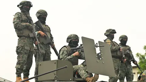 Five Ecuadorean soldiers stand atop an armoured vehicle at a command post in Guayas province. They are wearing camouflage uniforms, helmets and their faces are partly covered by black balaclavas. They are all heavily armed. 