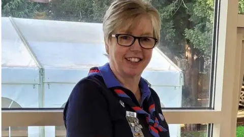 Girlguiding Anglia Tracy Foster wears a girl guides' uniform of cornflower blue shirt with a navy jumper and a twisted knotted red and cornflower blue scarf round her jumper.