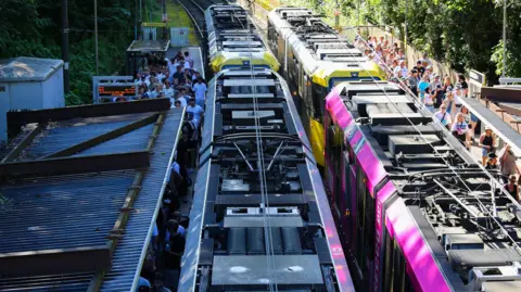 Emma Gibbs A long queue of Oasis fans stand under the corrugated iron roof of a Metrolink platform as a yellow and black liveried tram and a pink liveried tram stand at the stop