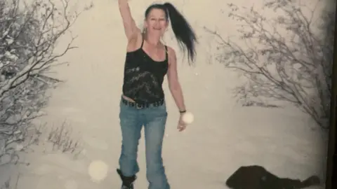 Stephen Alexander Woman standing in the snow with one hand in the air. She is wearing a strap top and jeans. Her dark hair is in a high ponytail.