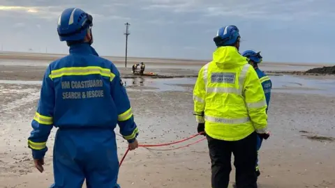 Wirral Coastguard Rescue Team Three members of HM Coastguard Search and Rescue, dressed in yellow and blue high visibility jackets, stand facing the muddy area of Leasowe Bay where a rescue is taking place on the beach.