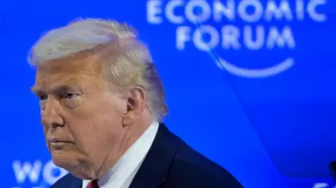 An image taken of Trump from his right side, showing his side profile as he looks away from the camera towards the left. He is wearing a suit with a red tie. Behind him is a blue screen with the World Economic Forum logo. 