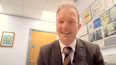 Tony McCabe is a middle-aged man with fair hair. He wears a brown suit, light shirt and stripey tie. He has a lanyard that reads 'staff'. There is a classroom display in the background.