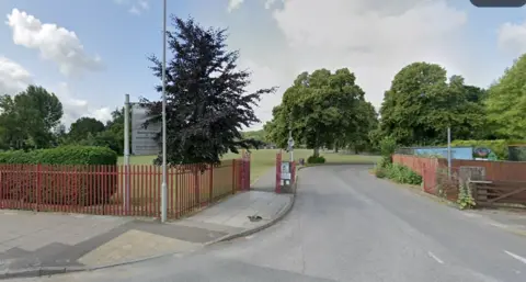 Fenced entrance with sign at school - there are tree-lined paths and glimpses of buildings.