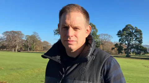 A man wearing a dark gillet , stood in front of parkland 