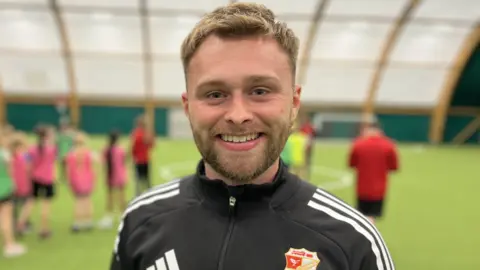 Andy Tye tersenyum dan mengenakan atasan Swindon Town FC Foundation berwarna hitam. Dia memiliki rambut pendek berwarna coklat tikus dan janggut.
