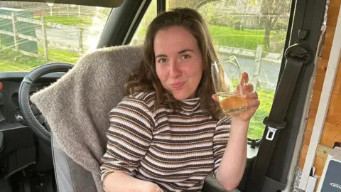 Handout A woman with mid-length brown hair sat inside a van drinking a glass of juice, eating her breakfast 